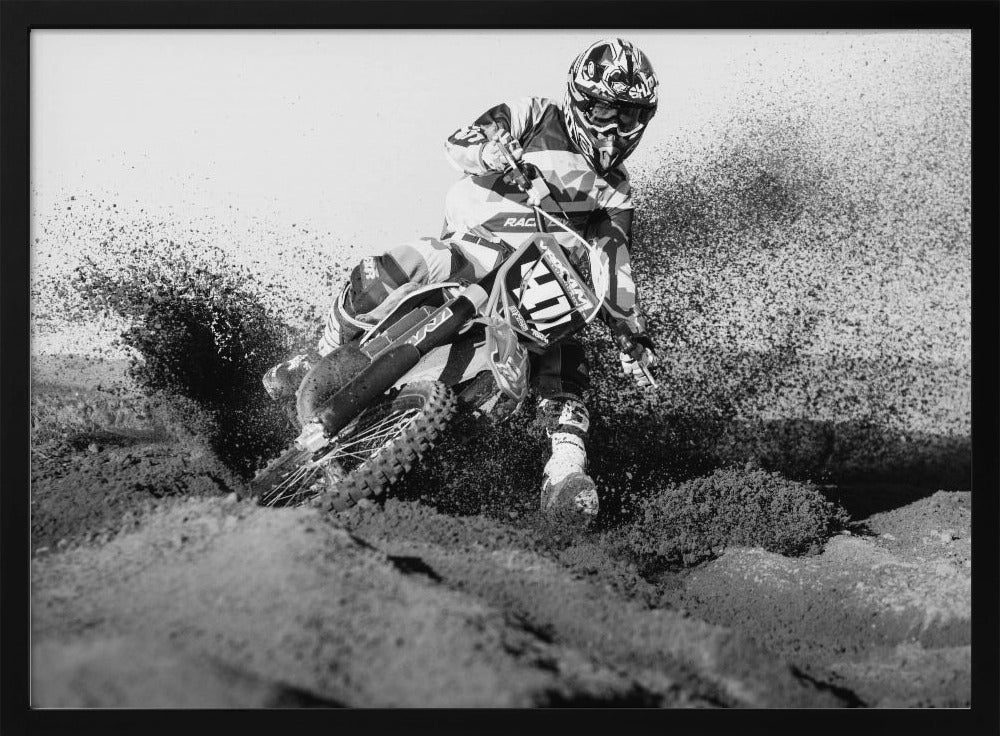 A dynamic black and white action photo of a motocross rider in full gear leaning hard into a turn, kicking up a large spray of dirt and sand from the track. The image is presented within a silver frame. Decor