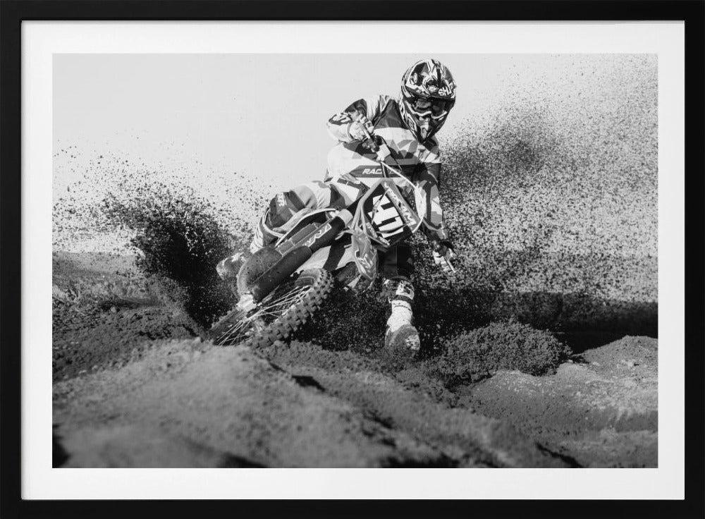 A dynamic black and white action photo of a motocross rider in full gear leaning hard into a turn, kicking up a large spray of dirt and sand from the track. The image is presented within a silver frame. Decor
