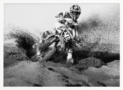 A dynamic black and white action photo of a motocross rider in full gear leaning hard into a turn, kicking up a large spray of dirt and sand from the track. The image is presented within a silver frame. Decor