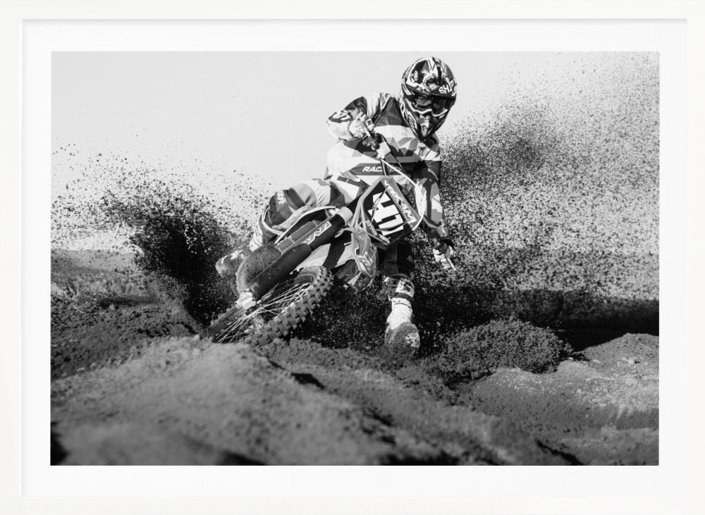 A dynamic black and white action photo of a motocross rider in full gear leaning hard into a turn, kicking up a large spray of dirt and sand from the track. The image is presented within a silver frame. Decor