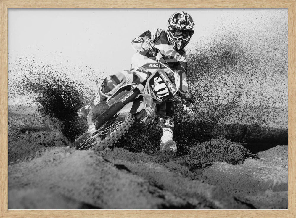 A dynamic black and white action photo of a motocross rider in full gear leaning hard into a turn, kicking up a large spray of dirt and sand from the track. The image is presented within a silver frame. Decor