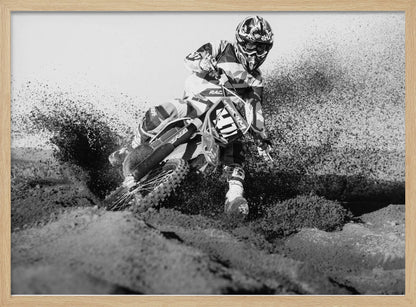 A dynamic black and white action photo of a motocross rider in full gear leaning hard into a turn, kicking up a large spray of dirt and sand from the track. The image is presented within a silver frame. Decor