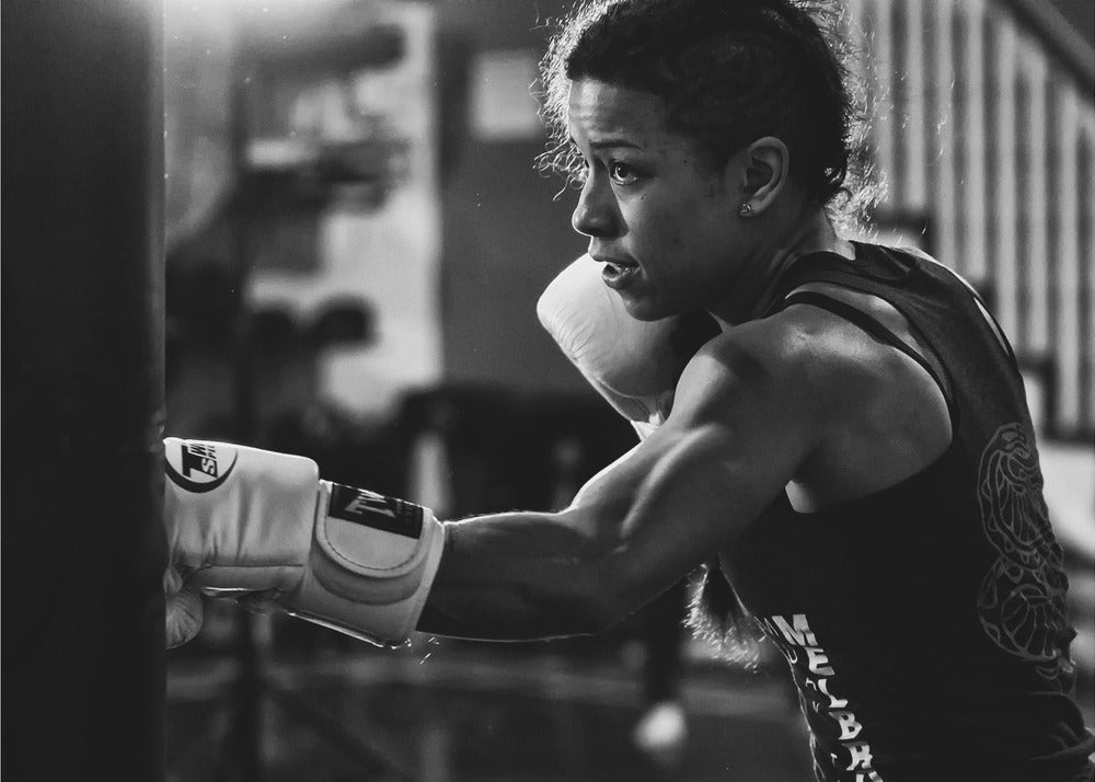 A dramatic black and white photo of a muscular female boxer in profile, intensely focused as she punches a heavy bag, set within a silver frame. Print