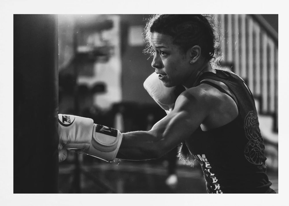 A dramatic black and white photo of a muscular female boxer in profile, intensely focused as she punches a heavy bag, set within a silver frame. Print