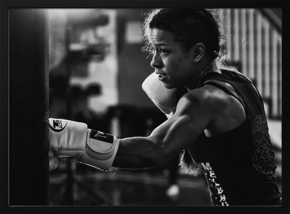A dramatic black and white photo of a muscular female boxer in profile, intensely focused as she punches a heavy bag, set within a silver frame. Print