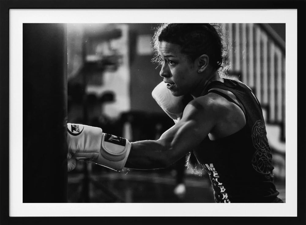 A dramatic black and white photo of a muscular female boxer in profile, intensely focused as she punches a heavy bag, set within a silver frame. Print