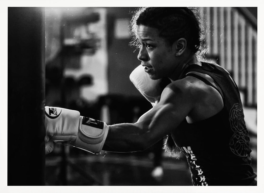 A dramatic black and white photo of a muscular female boxer in profile, intensely focused as she punches a heavy bag, set within a silver frame. Print