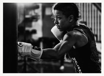 A dramatic black and white photo of a muscular female boxer in profile, intensely focused as she punches a heavy bag, set within a silver frame. Print