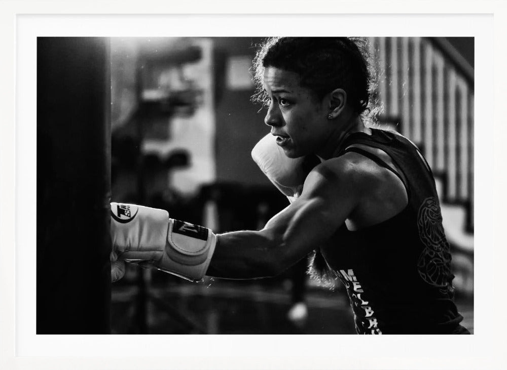 A dramatic black and white photo of a muscular female boxer in profile, intensely focused as she punches a heavy bag, set within a silver frame. Print