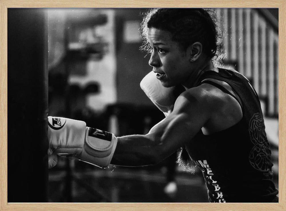 A dramatic black and white photo of a muscular female boxer in profile, intensely focused as she punches a heavy bag, set within a silver frame. Print