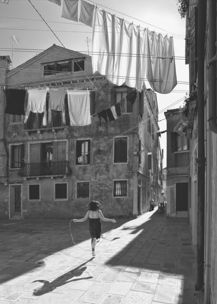 A black and white photograph capturing a girl from behind as she jump-ropes down a sunlit, narrow cobblestone street in Venice. Her long shadow is cast before her, and above, laundry hangs on clotheslines strung between old, weathered European buildings. Wall Art