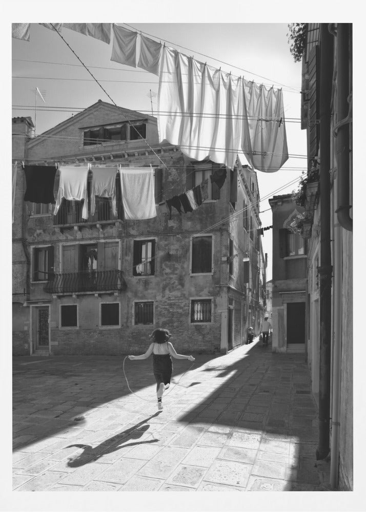 A black and white photograph capturing a girl from behind as she jump-ropes down a sunlit, narrow cobblestone street in Venice. Her long shadow is cast before her, and above, laundry hangs on clotheslines strung between old, weathered European buildings. Wall Art