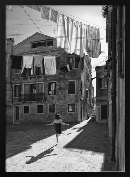 A black and white photograph capturing a girl from behind as she jump-ropes down a sunlit, narrow cobblestone street in Venice. Her long shadow is cast before her, and above, laundry hangs on clotheslines strung between old, weathered European buildings. Wall Art