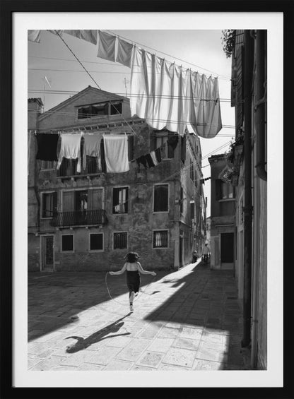 A black and white photograph capturing a girl from behind as she jump-ropes down a sunlit, narrow cobblestone street in Venice. Her long shadow is cast before her, and above, laundry hangs on clotheslines strung between old, weathered European buildings. Wall Art