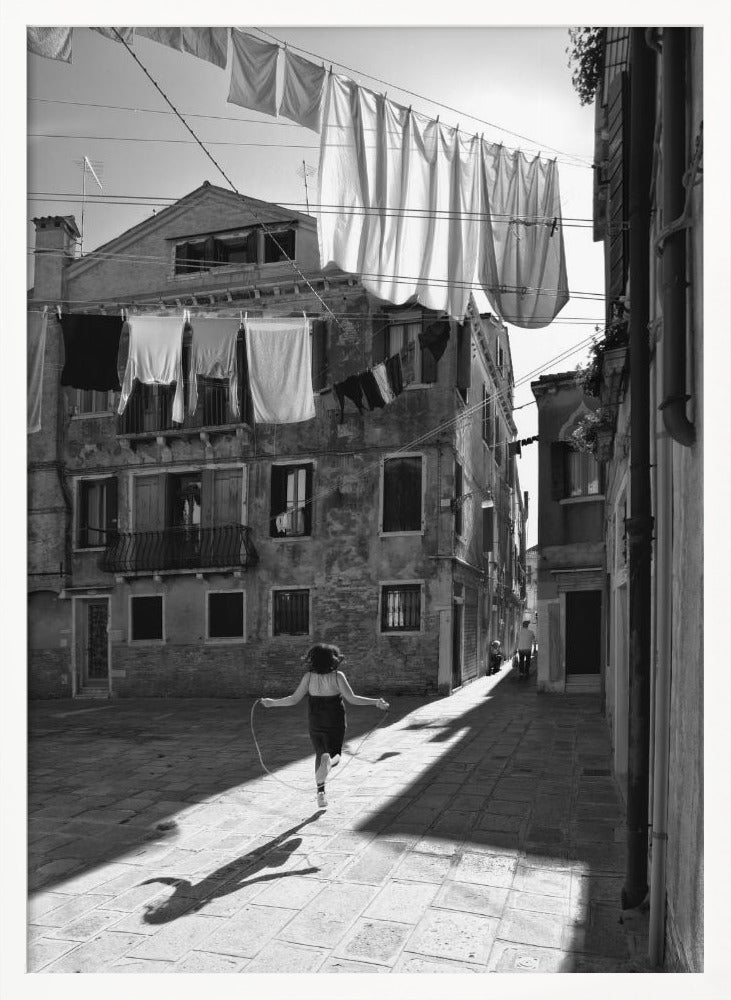 A black and white photograph capturing a girl from behind as she jump-ropes down a sunlit, narrow cobblestone street in Venice. Her long shadow is cast before her, and above, laundry hangs on clotheslines strung between old, weathered European buildings. Wall Art