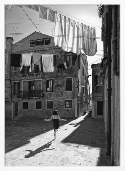 A black and white photograph capturing a girl from behind as she jump-ropes down a sunlit, narrow cobblestone street in Venice. Her long shadow is cast before her, and above, laundry hangs on clotheslines strung between old, weathered European buildings. Wall Art