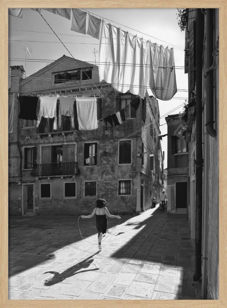 A black and white photograph capturing a girl from behind as she jump-ropes down a sunlit, narrow cobblestone street in Venice. Her long shadow is cast before her, and above, laundry hangs on clotheslines strung between old, weathered European buildings. Wall Art