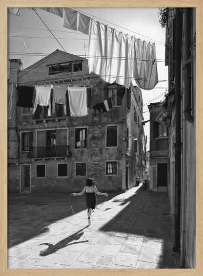 A black and white photograph capturing a girl from behind as she jump-ropes down a sunlit, narrow cobblestone street in Venice. Her long shadow is cast before her, and above, laundry hangs on clotheslines strung between old, weathered European buildings. Wall Art
