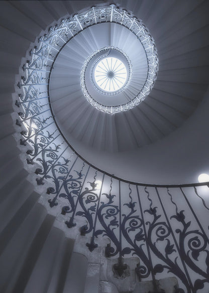A dramatic low-angle view looking up a majestic spiral staircase, its ornate black wrought iron railings creating a dark, intricate pattern against the cool-toned white steps that wind towards a circular skylight at the apex. Decor