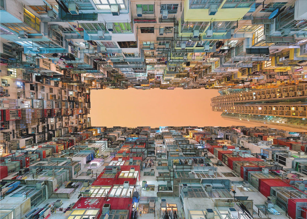 An upward-looking photograph from a courtyard surrounded by densely packed, multi-colored apartment buildings that create a rectangular frame around an orange sky. The unique perspective creates a surreal and geometric view of an urban landscape, all enclosed in a silver frame. Artwork