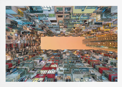 An upward-looking photograph from a courtyard surrounded by densely packed, multi-colored apartment buildings that create a rectangular frame around an orange sky. The unique perspective creates a surreal and geometric view of an urban landscape, all enclosed in a silver frame. Artwork