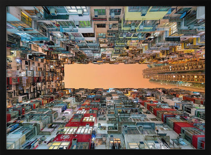 An upward-looking photograph from a courtyard surrounded by densely packed, multi-colored apartment buildings that create a rectangular frame around an orange sky. The unique perspective creates a surreal and geometric view of an urban landscape, all enclosed in a silver frame. Artwork