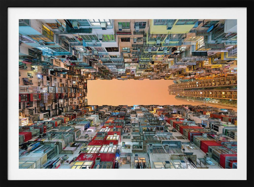 An upward-looking photograph from a courtyard surrounded by densely packed, multi-colored apartment buildings that create a rectangular frame around an orange sky. The unique perspective creates a surreal and geometric view of an urban landscape, all enclosed in a silver frame. Artwork