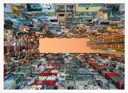 An upward-looking photograph from a courtyard surrounded by densely packed, multi-colored apartment buildings that create a rectangular frame around an orange sky. The unique perspective creates a surreal and geometric view of an urban landscape, all enclosed in a silver frame. Artwork