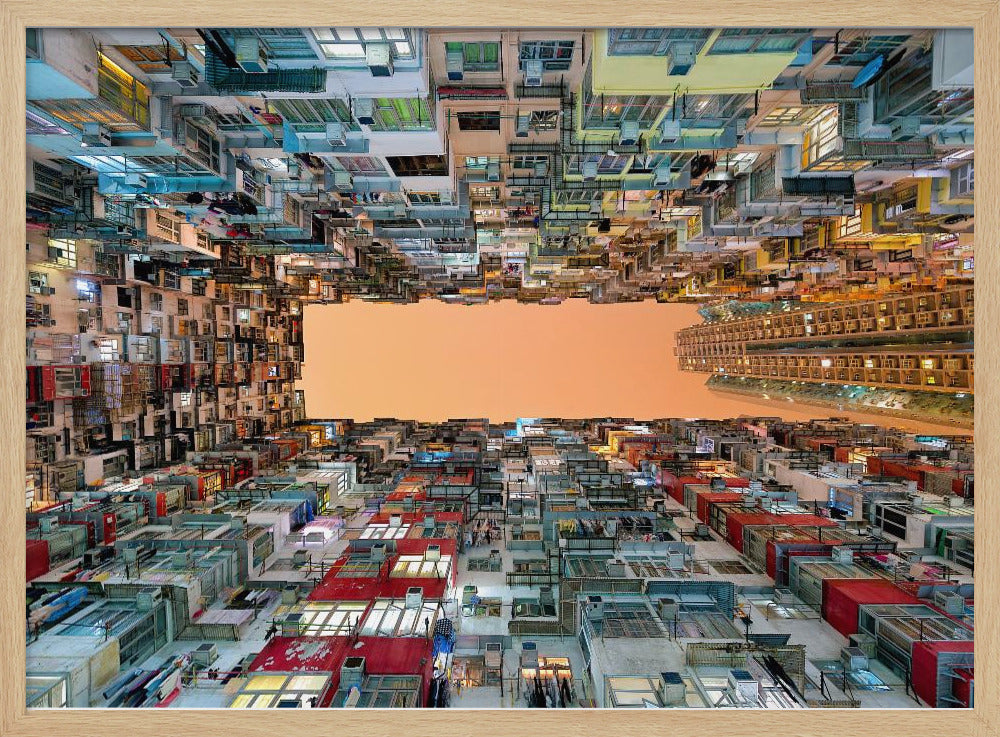 An upward-looking photograph from a courtyard surrounded by densely packed, multi-colored apartment buildings that create a rectangular frame around an orange sky. The unique perspective creates a surreal and geometric view of an urban landscape, all enclosed in a silver frame. Artwork