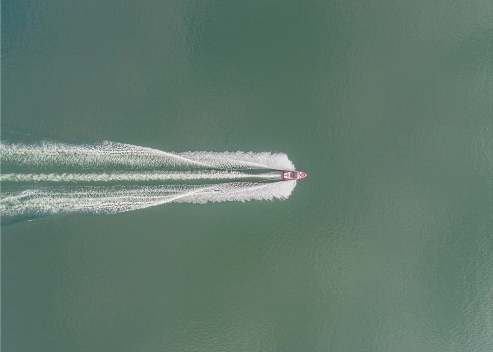An aerial top-down photograph of a red motorboat speeding across a vast expanse of calm, green water, leaving a long, white, frothy wake behind it. The scene is presented within a simple silver frame. Print