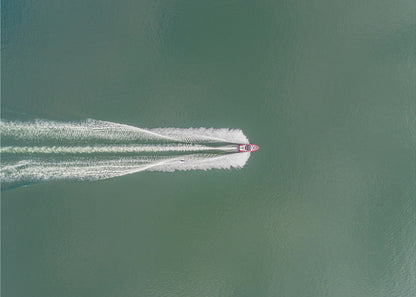 An aerial top-down photograph of a red motorboat speeding across a vast expanse of calm, green water, leaving a long, white, frothy wake behind it. The scene is presented within a simple silver frame. Print