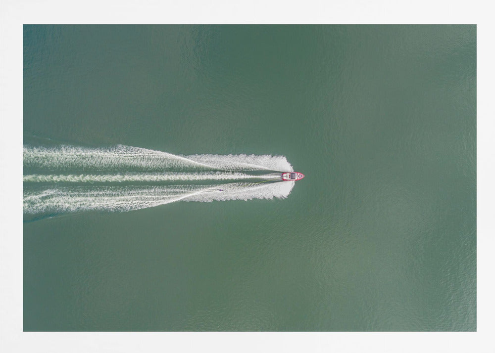 An aerial top-down photograph of a red motorboat speeding across a vast expanse of calm, green water, leaving a long, white, frothy wake behind it. The scene is presented within a simple silver frame. Print