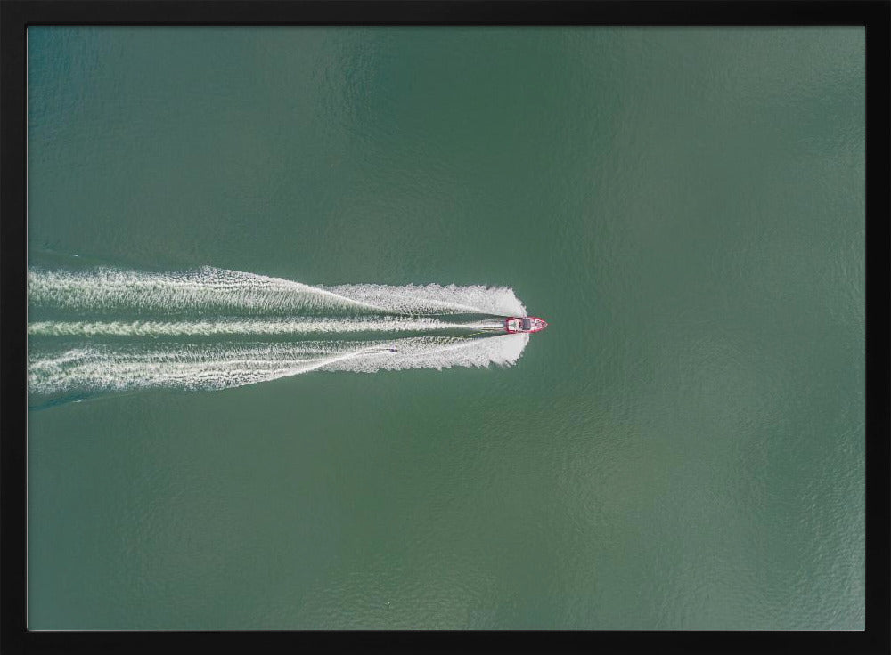 An aerial top-down photograph of a red motorboat speeding across a vast expanse of calm, green water, leaving a long, white, frothy wake behind it. The scene is presented within a simple silver frame. Print