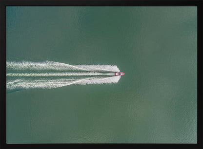 An aerial top-down photograph of a red motorboat speeding across a vast expanse of calm, green water, leaving a long, white, frothy wake behind it. The scene is presented within a simple silver frame. Print