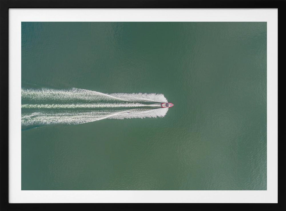 An aerial top-down photograph of a red motorboat speeding across a vast expanse of calm, green water, leaving a long, white, frothy wake behind it. The scene is presented within a simple silver frame. Print