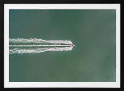 An aerial top-down photograph of a red motorboat speeding across a vast expanse of calm, green water, leaving a long, white, frothy wake behind it. The scene is presented within a simple silver frame. Print