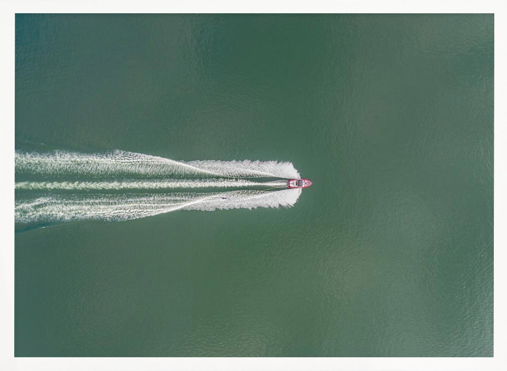 An aerial top-down photograph of a red motorboat speeding across a vast expanse of calm, green water, leaving a long, white, frothy wake behind it. The scene is presented within a simple silver frame. Print