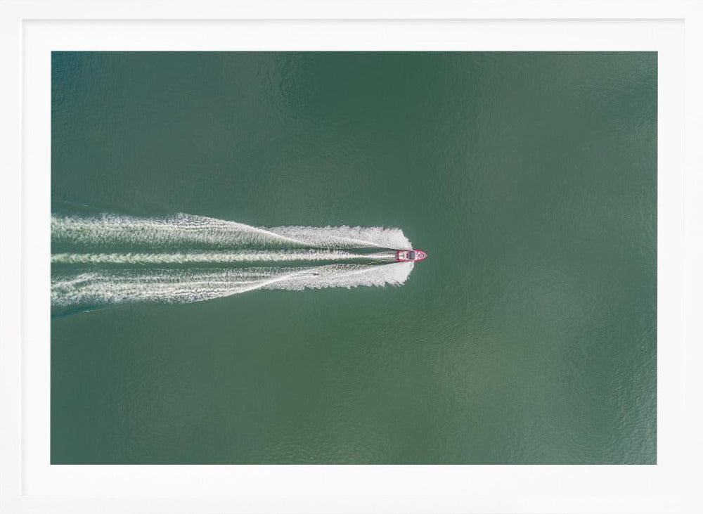 An aerial top-down photograph of a red motorboat speeding across a vast expanse of calm, green water, leaving a long, white, frothy wake behind it. The scene is presented within a simple silver frame. Print