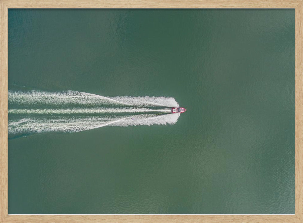 An aerial top-down photograph of a red motorboat speeding across a vast expanse of calm, green water, leaving a long, white, frothy wake behind it. The scene is presented within a simple silver frame. Print