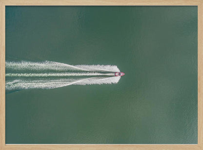 An aerial top-down photograph of a red motorboat speeding across a vast expanse of calm, green water, leaving a long, white, frothy wake behind it. The scene is presented within a simple silver frame. Print