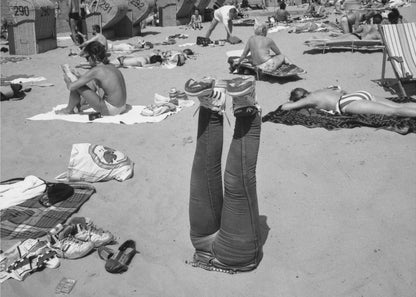 A quirky, vintage black and white photograph of a crowded beach. The central focus is a person lying on their back with their legs straight up in the air, oddly wearing jeans and sneakers. Surrounding them are numerous other beachgoers sunbathing on towels and in wicker beach chairs, creating a busy and candid summer scene. Wall Art