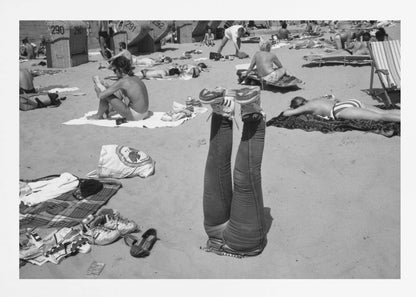 A quirky, vintage black and white photograph of a crowded beach. The central focus is a person lying on their back with their legs straight up in the air, oddly wearing jeans and sneakers. Surrounding them are numerous other beachgoers sunbathing on towels and in wicker beach chairs, creating a busy and candid summer scene. Wall Art