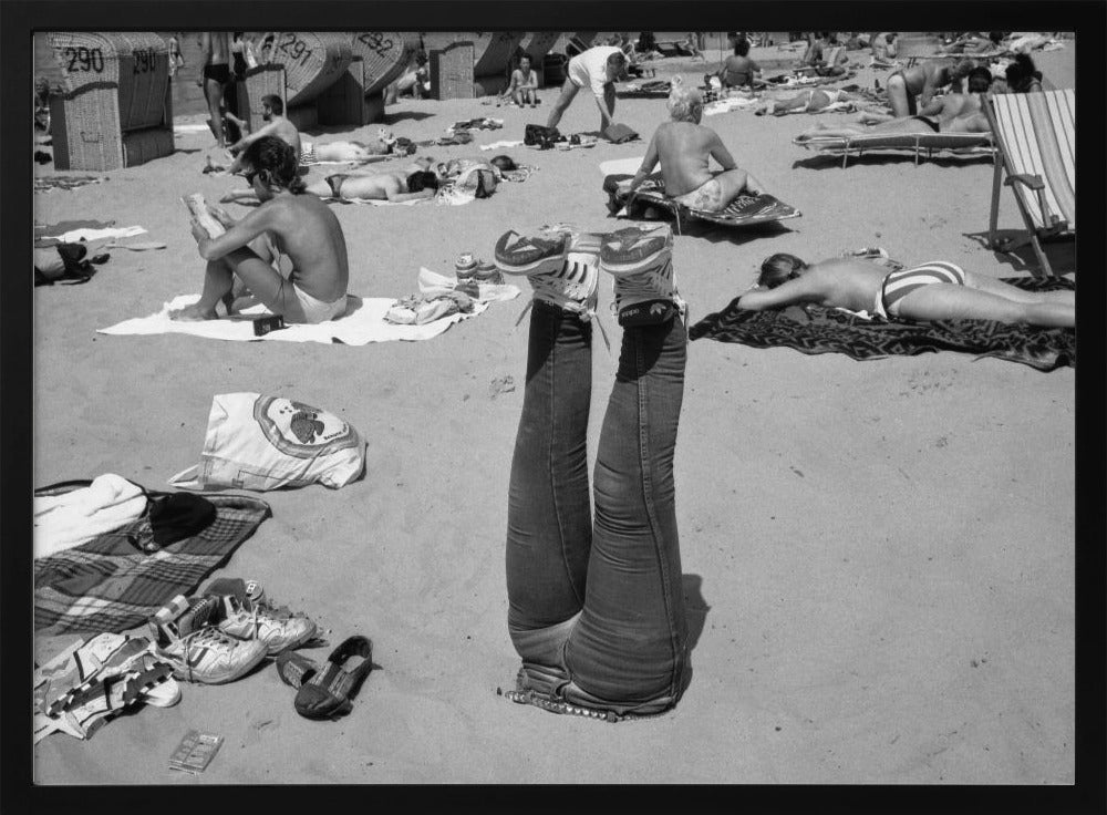 A quirky, vintage black and white photograph of a crowded beach. The central focus is a person lying on their back with their legs straight up in the air, oddly wearing jeans and sneakers. Surrounding them are numerous other beachgoers sunbathing on towels and in wicker beach chairs, creating a busy and candid summer scene. Wall Art
