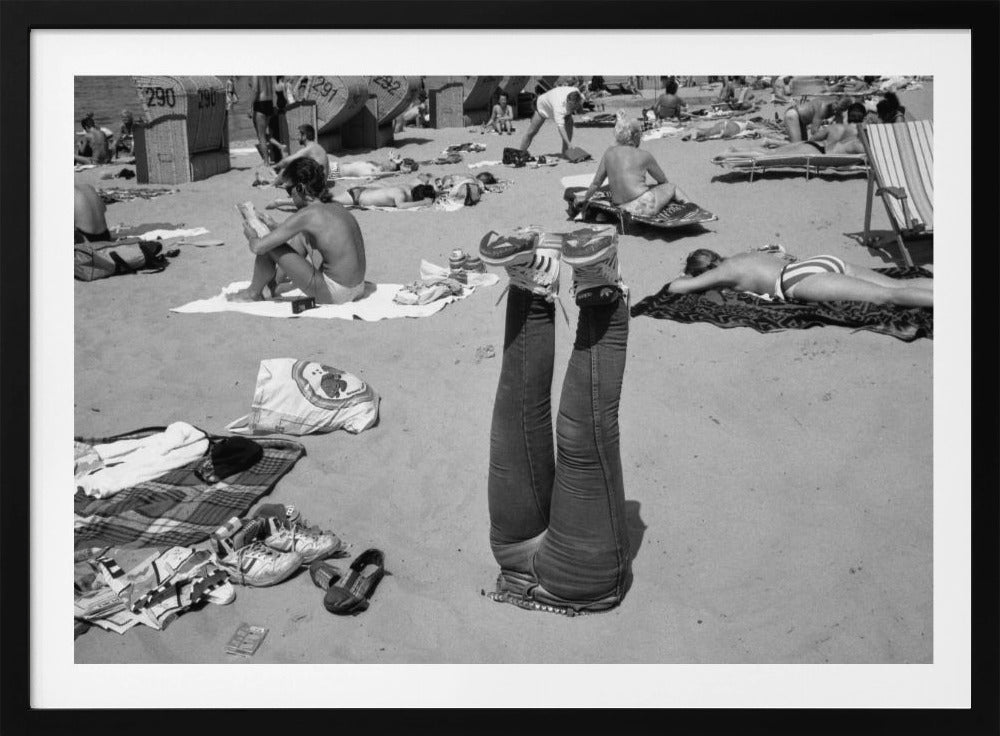 A quirky, vintage black and white photograph of a crowded beach. The central focus is a person lying on their back with their legs straight up in the air, oddly wearing jeans and sneakers. Surrounding them are numerous other beachgoers sunbathing on towels and in wicker beach chairs, creating a busy and candid summer scene. Wall Art
