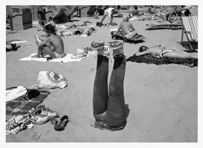 A quirky, vintage black and white photograph of a crowded beach. The central focus is a person lying on their back with their legs straight up in the air, oddly wearing jeans and sneakers. Surrounding them are numerous other beachgoers sunbathing on towels and in wicker beach chairs, creating a busy and candid summer scene. Wall Art