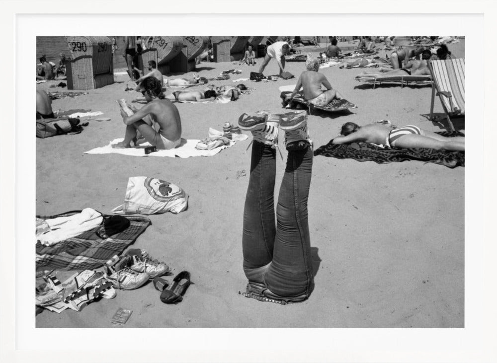 A quirky, vintage black and white photograph of a crowded beach. The central focus is a person lying on their back with their legs straight up in the air, oddly wearing jeans and sneakers. Surrounding them are numerous other beachgoers sunbathing on towels and in wicker beach chairs, creating a busy and candid summer scene. Wall Art