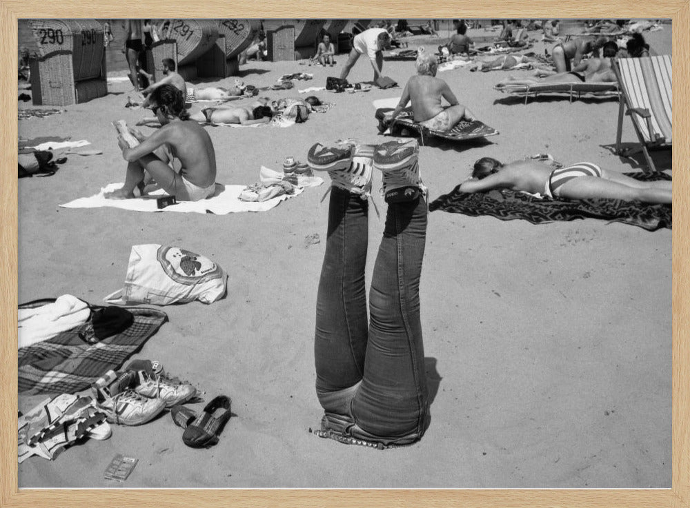 A quirky, vintage black and white photograph of a crowded beach. The central focus is a person lying on their back with their legs straight up in the air, oddly wearing jeans and sneakers. Surrounding them are numerous other beachgoers sunbathing on towels and in wicker beach chairs, creating a busy and candid summer scene. Wall Art