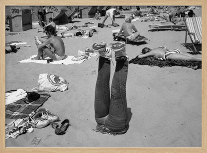 A quirky, vintage black and white photograph of a crowded beach. The central focus is a person lying on their back with their legs straight up in the air, oddly wearing jeans and sneakers. Surrounding them are numerous other beachgoers sunbathing on towels and in wicker beach chairs, creating a busy and candid summer scene. Wall Art
