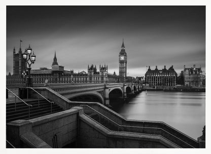 A framed, black and white, long-exposure photograph of the London skyline featuring the Houses of Parliament and the Big Ben clock tower across the River Thames. In the foreground, stone steps lead down towards Westminster Bridge, and the water of the river is smooth and reflective under a dark, cloudy sky. Print