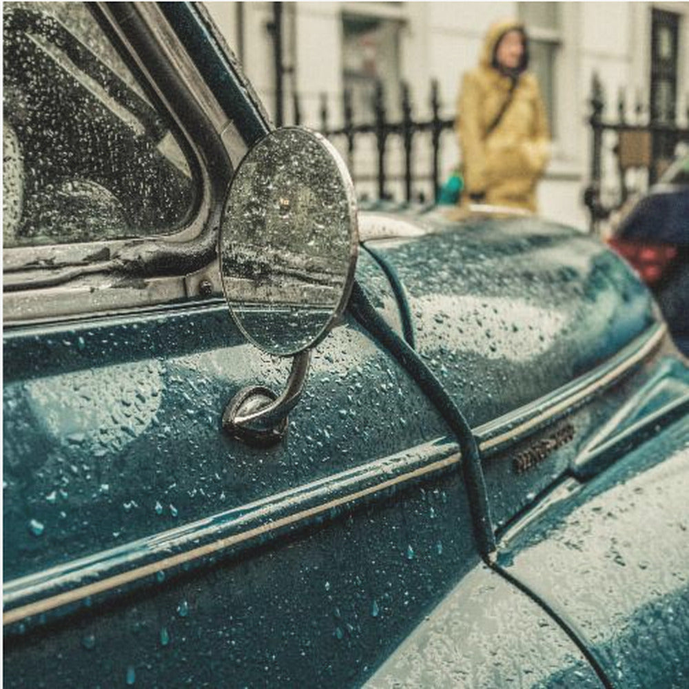 A close-up, moody photograph of a vintage teal car on a rainy day, with raindrops covering its surface. The oval side-view mirror reflects the wet street, and in the background, a person in a yellow raincoat is visible. Wall Art
